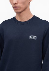 Homme portant un pull col rond bleu foncé avec un écusson à logo EA7 Emporio Armani sur la poitrine gauche.