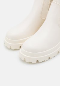 Bottines blanches avec finition en cuir lisse, semelle épaisse blanche, bout arrondi et panneaux latéraux élastiques pour un enfilage facile.