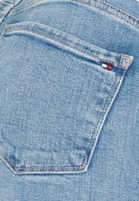 Tela de mezclilla azul claro con un bolsillo trasero distintivo; presenta una pequeña etiqueta de logo en rojo, blanco y azul en el borde del bolsillo.