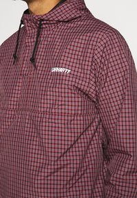 Rood en marineblauw geblokte hooded jack met een half-zip voorkant, voorzien van een logo en verstelbare trekkoorden aan de capuchon en de onderzijde.