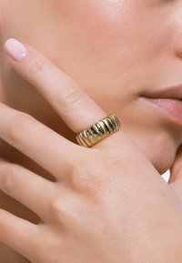 Gouden ring met een gestructureerde band met horizontale strepen, liggend op een licht gebruinde hand, met een lichtroze nagellak zichtbaar.