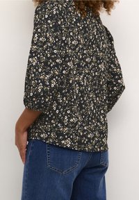 Sort blomsterbluse med trekvart ærmer, med en blanding af hvide og gule blomster på et tekstureret stof. Kombineret med blå denimjeans.