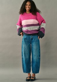Rosa och grå randig tröja med vida ärmar, kombinerad med ljus tvättade baggy jeans och svarta ballerinas, på en modell mot en neutral bakgrund.