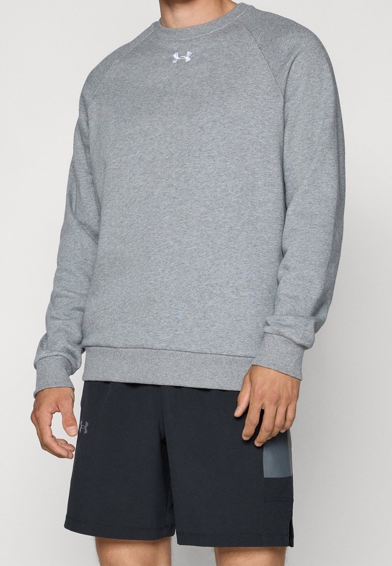 Homme portant un sweat-shirt gris à manches longues avec un logo blanc sur la poitrine et un short de sport noir avec une poche latérale.