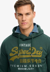 Jonge man draagt een donkergroene SuperDry-hoodie met gele en witte tekst, gecombineerd met een spijkerblouse met kraag.