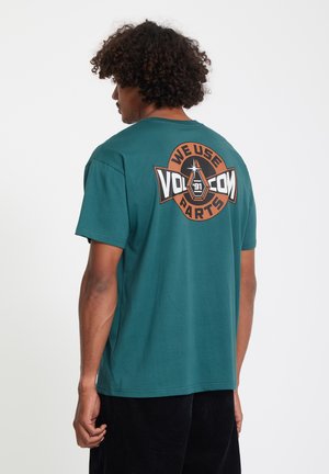 Man met krullend haar draagt een teal Volcom T-shirt met een ronde oranje-zwart logo waarop staat "We use Volcom parts '91" op de achterkant.