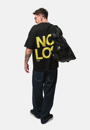 Mężczyzna w czarnej oversize koszulce z żółtym napisem "NO LOS", ciemnych dżinsach, czarnych tenisówkach, niosący dużą czarną torbę na jednym ramieniu.
