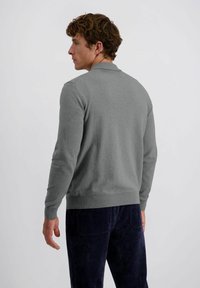 Man in grijze lange mouwen trui en donkerblauwe corduroy broek, staande met het gezicht naar links tegen een eenvoudige lichtgrijze achtergrond.