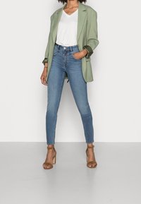 Blazer verde claro sobre una camiseta blanca con escote en V, combinado con jeans skinny azules y sandalias de tacón color marrón. Tejido suave y corte ajustado.