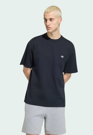 Junger Mann mit kurzen blonden Haaren, trägt ein schwarzes Adidas-Logo-T-Shirt und hellgraue Shorts, steht mit den Händen hinter dem Rücken.
