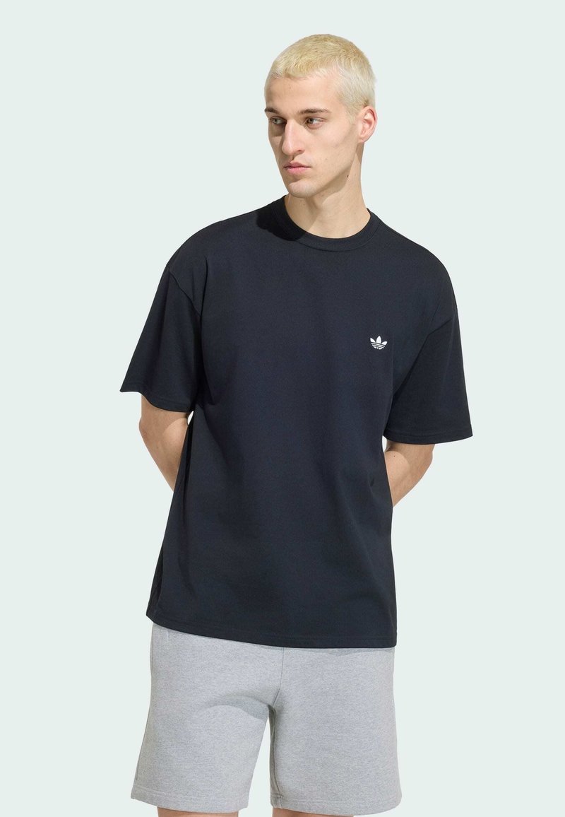 Jeune homme aux cheveux blonds courts portant un t-shirt noir avec le logo Adidas et un short gris clair, debout les mains dans le dos.