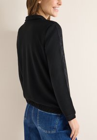 Giacca sportiva nera con colletto e branding discreto sulle maniche; presenta una vita aderente e una texture liscia. Abbinata a jeans in denim.