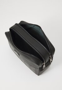 Svart lærveske med teksturert side, doble glidelåser og glatt finish. Innvendig viser en ensfarget mørk farge, designet for kompakt lagring.
