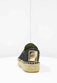 Espadrille de lona negra con superficie texturizada, suela de yute y una placa de logo en tono dorado en el talón. Forma redondeada y parte trasera abierta.