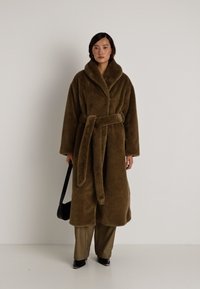 Casaco longo de pelo sintético castanho com uma gola larga, cintura cingida e mangas largas. Modelo combina com calças beiges e uma bolsa preta.