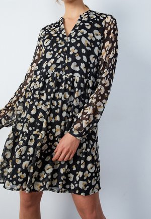Kobieta ubrana w czarną sukienkę z beżowymi i białymi abstrakcyjnymi wzorami kwiatowymi, z długimi przejrzystymi rękawami, guzikami przy dekolcie oraz spódnicą warstwową.