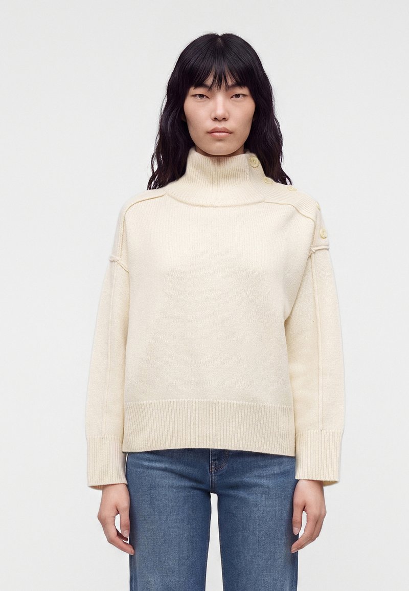 Femme aux longs cheveux noirs portant un pull à col roulé crème avec des boutons sur l’épaule et un jean bleu, debout devant un fond uni.