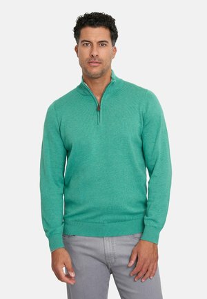 Suéter verde de punto con cuello con cremallera, que presenta un patrón texturizado. Mangas largas con puños acanalados y dobladillo, usado sobre pantalones grises.