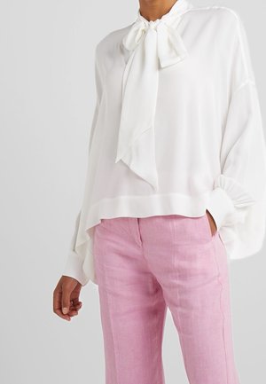 Osoba ubrana w luźną białą bluzkę z kołnierzykiem wiązanym na kokardę i długimi rękawami, w połączeniu z różowymi lnianymi spodniami z wysokim stanem na jednolitym tle.