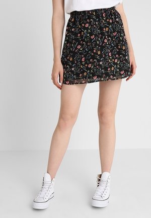 Femme portant une mini-jupe noire à motifs floraux associée à des baskets montantes blanches, debout devant un fond gris clair uni.