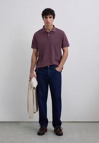 Camisa polo burdeos con cuello, jeans azul oscuro y zapatos marrones. El modelo sostiene un suéter crema. Diseño simple y limpio con un corte relajado.