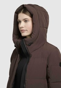 Giubbotto puffer marrone con un ampio cappuccio, chiusura con zip e trapuntatura visibile. Presenta accessori neri e branding sulla zip.