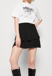 Žena v bílé grafické tričko zastrčeném do černé patrové mini sukně, s černými kozačkami nad kolena a stříbrným prstenem.