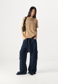 Beige medvilnės marškinėliai su balta "TOMMY HILFIGER" logotipu. Mėlynos plačios kojų kargo džinsai su kišenėmis ir diržu. Modelis pozuoja su petine krepšiu.