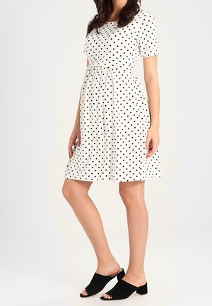 Femme portant une robe blanche à pois noirs longueur genoux et des mules à talons ouvertes noires, debout les mains détendues le long du corps.