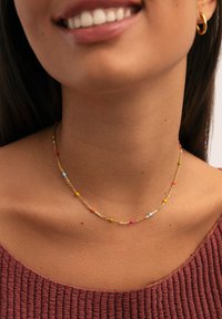 Una donna sorridente indossa una collana d'oro con piccole perline colorate e un orecchino a cerchio d'oro, vestita con un top rosso-bruno a texture.