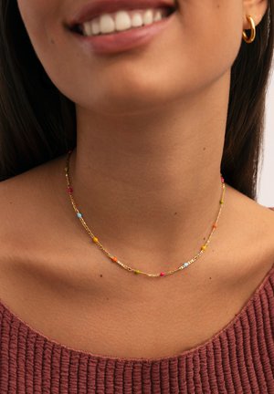 Una donna sorridente indossa una collana d'oro con piccole perline colorate e un orecchino a cerchio d'oro, vestita con un top rosso-bruno a texture.