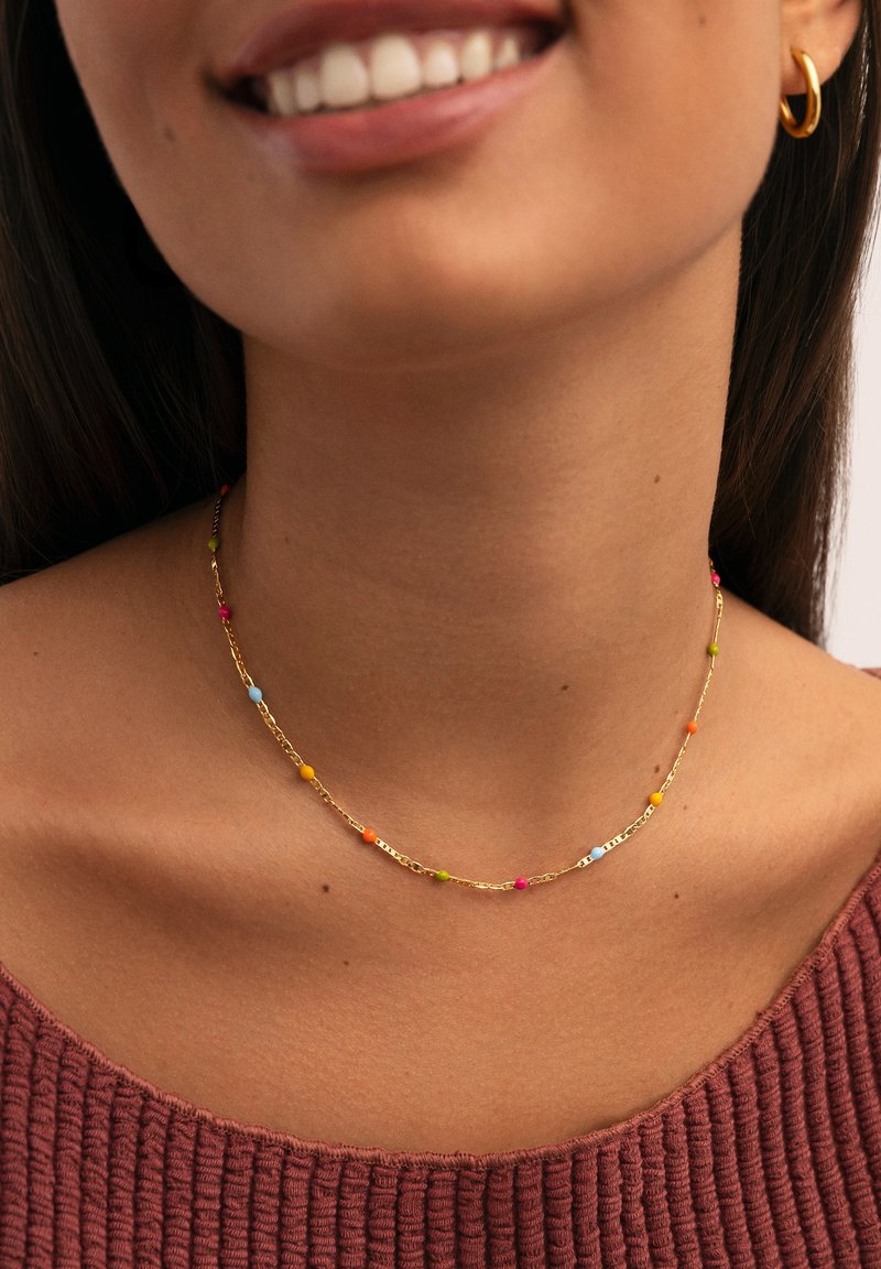 Una donna sorridente indossa una collana d'oro con piccole perline colorate e un orecchino a cerchio d'oro, vestita con un top rosso-bruno a texture.