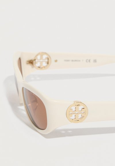 Lunettes de soleil couleur crème avec des verres marron et des accents de logo ronds en or sur les branches, présentées sur un fond blanc.