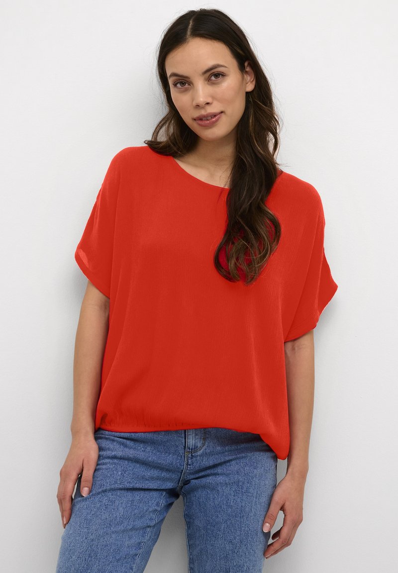 Blusa rossa fluente con scollatura ampia e maniche corte, abbinata a jeans azzurri; texture morbida, vestibilità comoda.