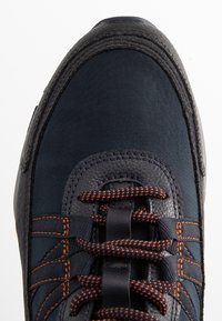 Zapatilla deportiva de color azul oscuro con material texturizado, costuras naranjas y cordones. Presenta una puntera reforzada y un collar acolchado para mayor soporte.