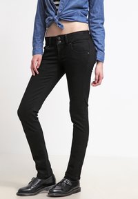 Pantalones skinny negros de cintura alta, con detalles de botones metálicos, bolsillos frontales y una textura suave, combinados con una blusa de denim.