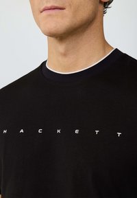 Zbliżenie na mężczyznę w czarnej koszulce z okrągłym dekoltem, z białymi literami rozłożonymi na piersi, tworzącymi napis "HACKETT".
