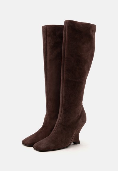 Bottes hautes en daim marron foncé avec un bout carré arrondi et un talon compensé courbé, debout sur un fond clair uni.