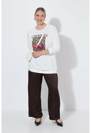 Donna sorridente con capelli chiari che indossa una felpa bianca con motivo di ciliegie, pantaloni larghi scuri, scarpe nere e una borsa bianca a tracolla.