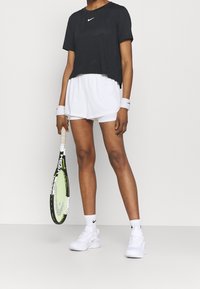 Camiseta negra corta con el logo blanco de Nike, pantalones cortos deportivos blancos, zapatillas blancas y sosteniendo una raqueta de tenis verde y negra.
