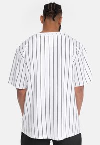 Hvid kortærmet t-shirt med sorte lodrette striber, der har en afslappet pasform og rund halsudskæring, set bagfra.