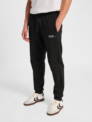 Uomo con pantaloni da jogging neri firmati Hummel e sneaker bianche con dettagli neri e marroni, in piedi con una mano in tasca.