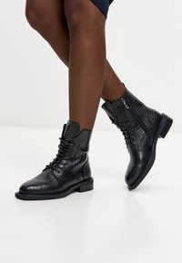 Botine din piele cu textură de crocodil, negre, cu șireturi și fermoare laterale, așezate peste picioare încrucișate, pe un fundal alb.