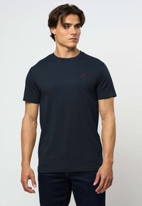 Muškarac koji nosi običnu tamnoplavu majicu kratkih rukava s okruglim izrezom i malim crvenim logotipom na prsima, stojeći nasuprot jednobojnoj svijetloj pozadini.