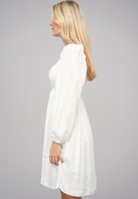 Vestido blanco con mangas largas abullonadas, tela texturada y un corpiño ajustado. La falda se ensancha suavemente, alcanzando la mitad del muslo en longitud.