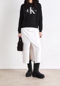 Černá mikina s kapucí a bílým logem, bílá midi sukně s předním rozparkem, černé kozačky nad kolena a malá hnědá texturovaná kabelka.