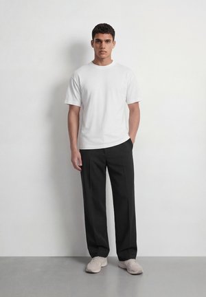 Jeune homme debout contre un mur blanc, portant une chemise blanche à manches courtes, un pantalon noir et des baskets de couleur claire, avec une main dans la poche.