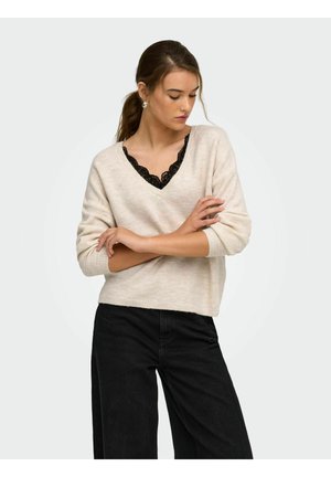 Femme aux cheveux foncés en queue de cheval, portant un pull beige à col V par-dessus un haut en dentelle noire et un pantalon noir, regardant vers le bas les bras croisés.