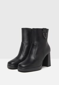 Schwarze Leder-Stiefeletten mit hohem Blockabsatz, Reißverschlussschluss und Kettenakzent an der Seite. Glatte Textur mit minimalen Nähdetails.