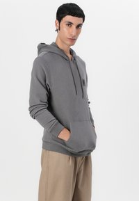 Moletão cinza com bolso frontal tipo canguru, capuz com cordão e detalhe de logotipo. Tecido macio e ajuste descontraído, combinado com calças bege.